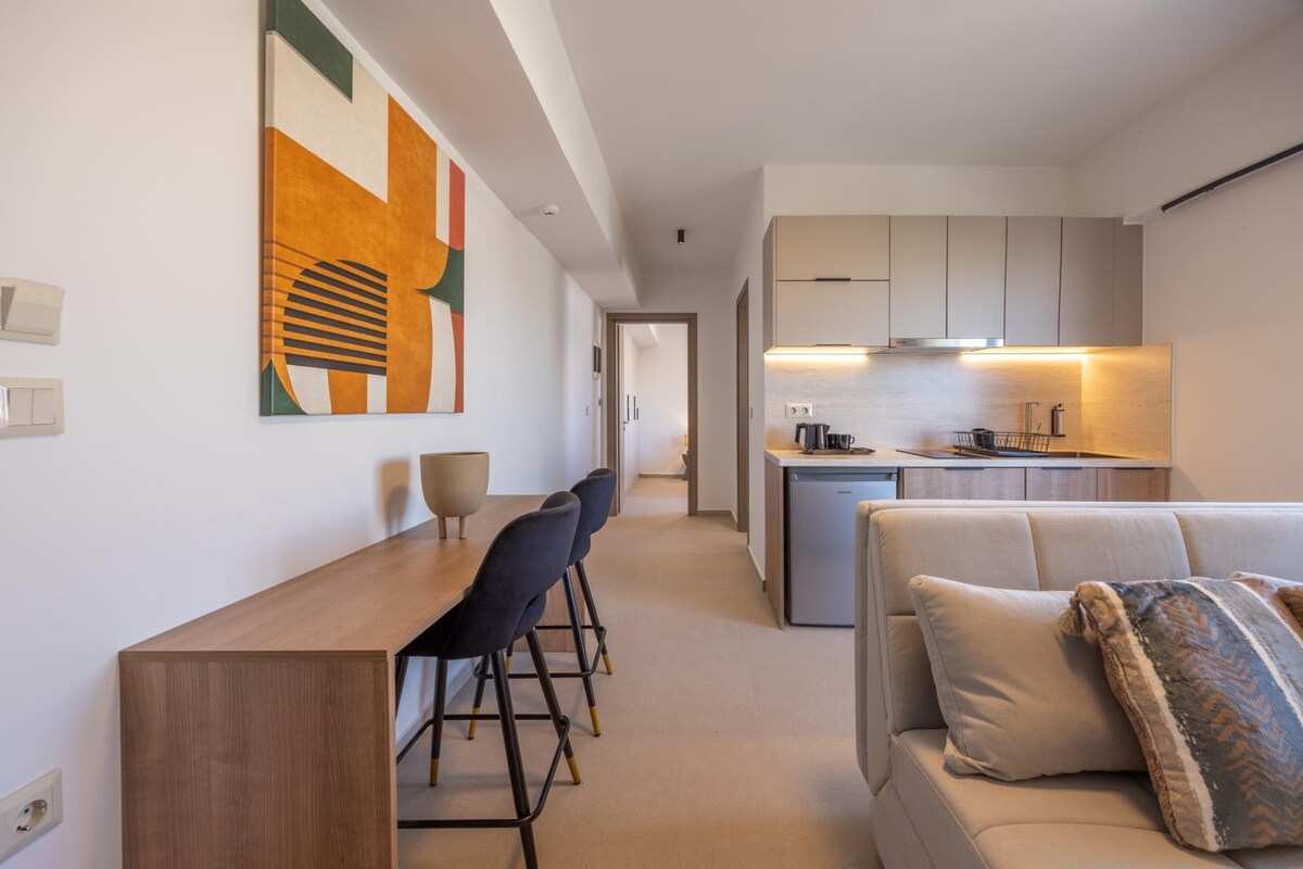 A modern kitchen with a central island, a dining area with a wooden table and chairs, and a living area with a sofa and cushions.