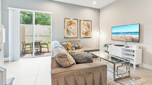 A modern living room with a sofa, coffee table, and TV. There are two framed artworks on the wall and a sliding glass door leading to an outdoor area. - Contemporary Summerville resort home - 5 miles to Disney (Kissimmee)