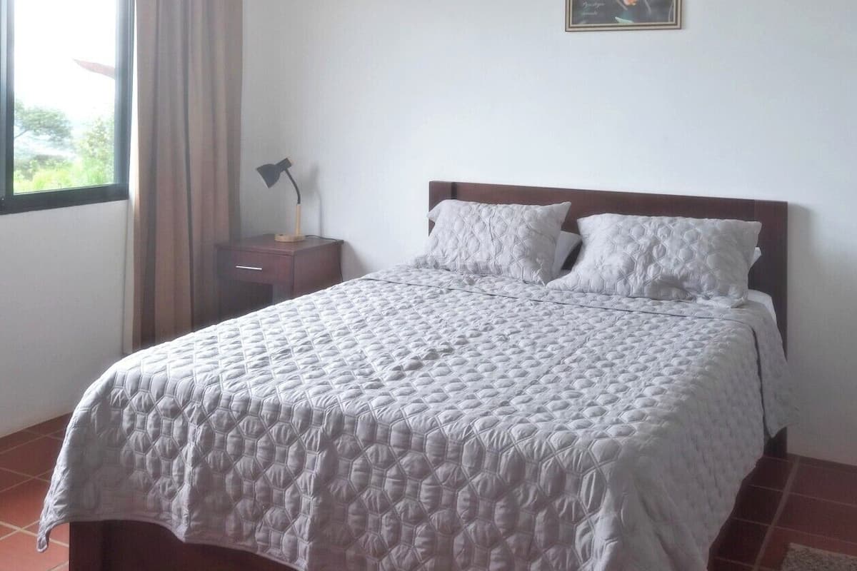 Een slaapkamer met een groot bed, een nachtkastje, een raam met gordijnen en een lijst met een afbeelding aan de muur.
