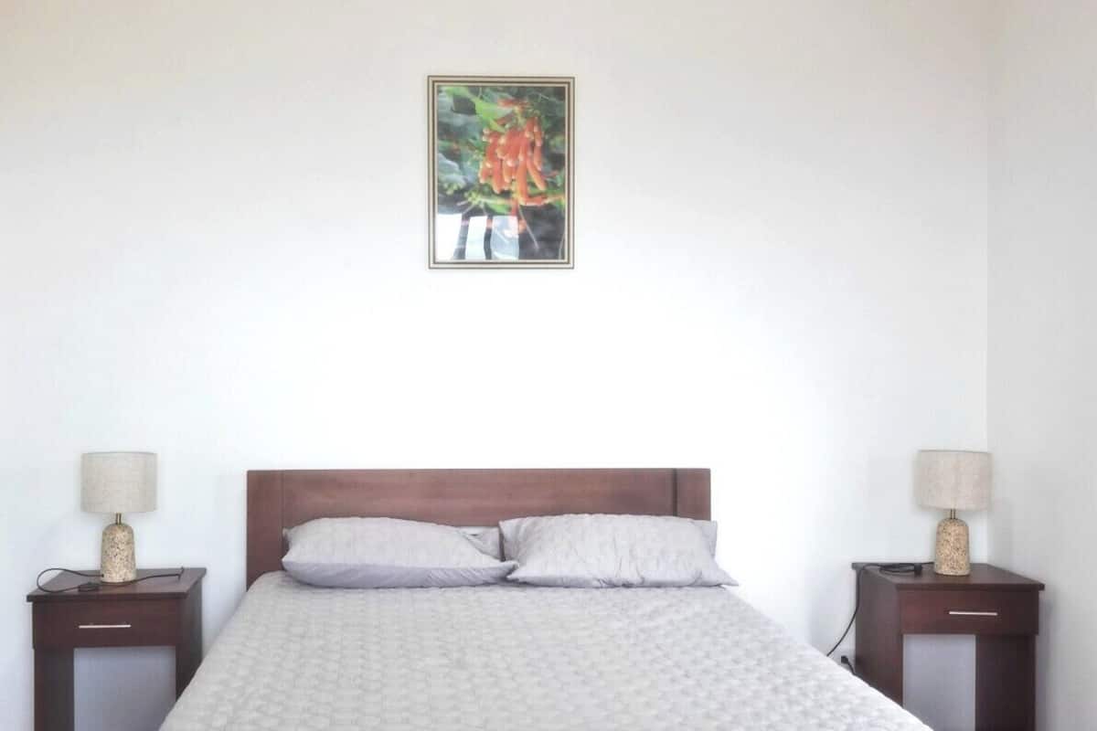 Een netjes opgemaakt bed met een witte quilt, twee nachtkastjes met lampen en een ingelijste foto aan de muur.