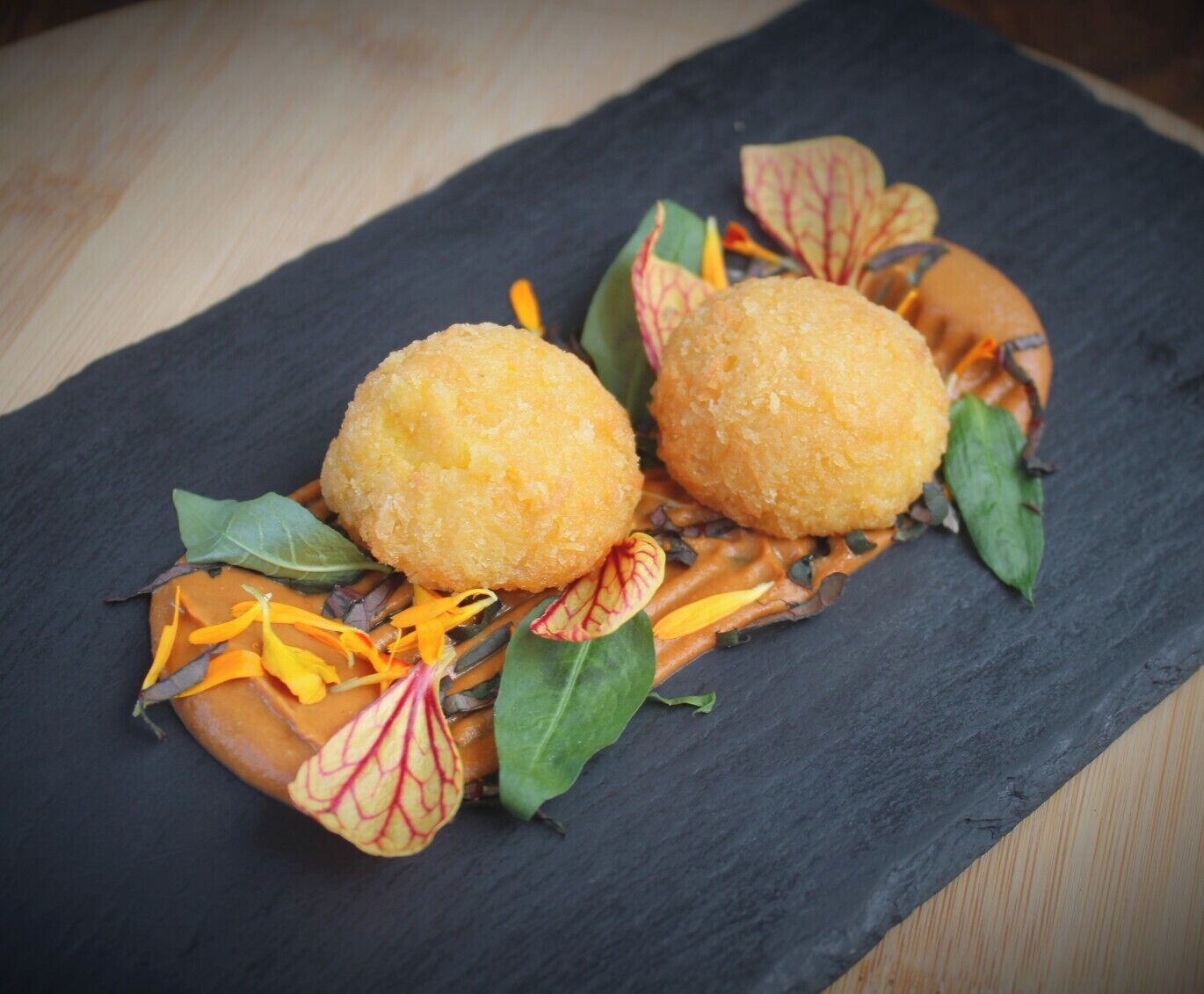 Two round, golden-brown fried items on a dark slate plate, garnished with green leaves and orange zest.