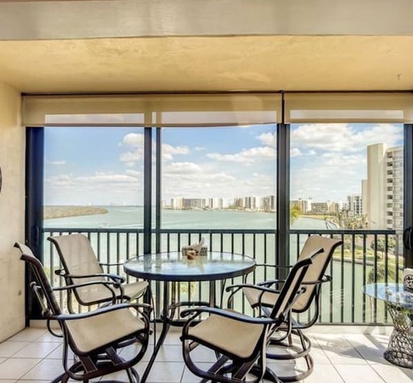 Un balcón con una mesa de vidrio y cuatro sillas, con vistas a un paisaje urbano y una masa de agua.