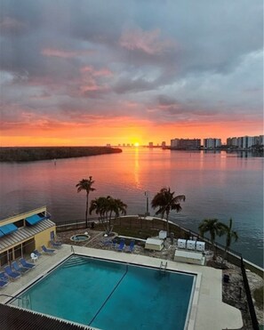 Een zwembad van een resort met uitzicht op een zonsondergang, palmbomen en een stadsachtergrond op de verte.