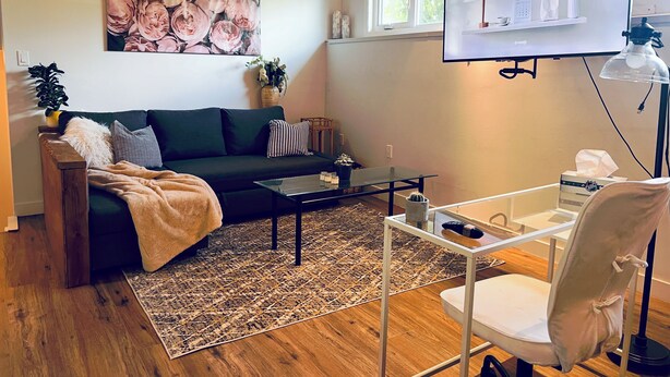 A modern living room with a dark green sofa, a glass coffee table, and a patterned rug. There's a TV mounted on the wall, a potted plant, and a cactus on a side table.