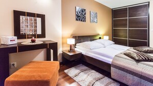 A hotel room with a bed, bedside table, lamp, radio, artwork, and a small orange ottoman.