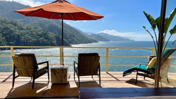 Een balkon met uitzicht op zee en bergen, twee stoelen, een kleine tafel en een rode parasol.