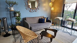 A modern living room with a grey sofa, a coffee table, a wicker chair, and a floor lamp. There is a large mirror on the wall and a potted plant on a shelf.