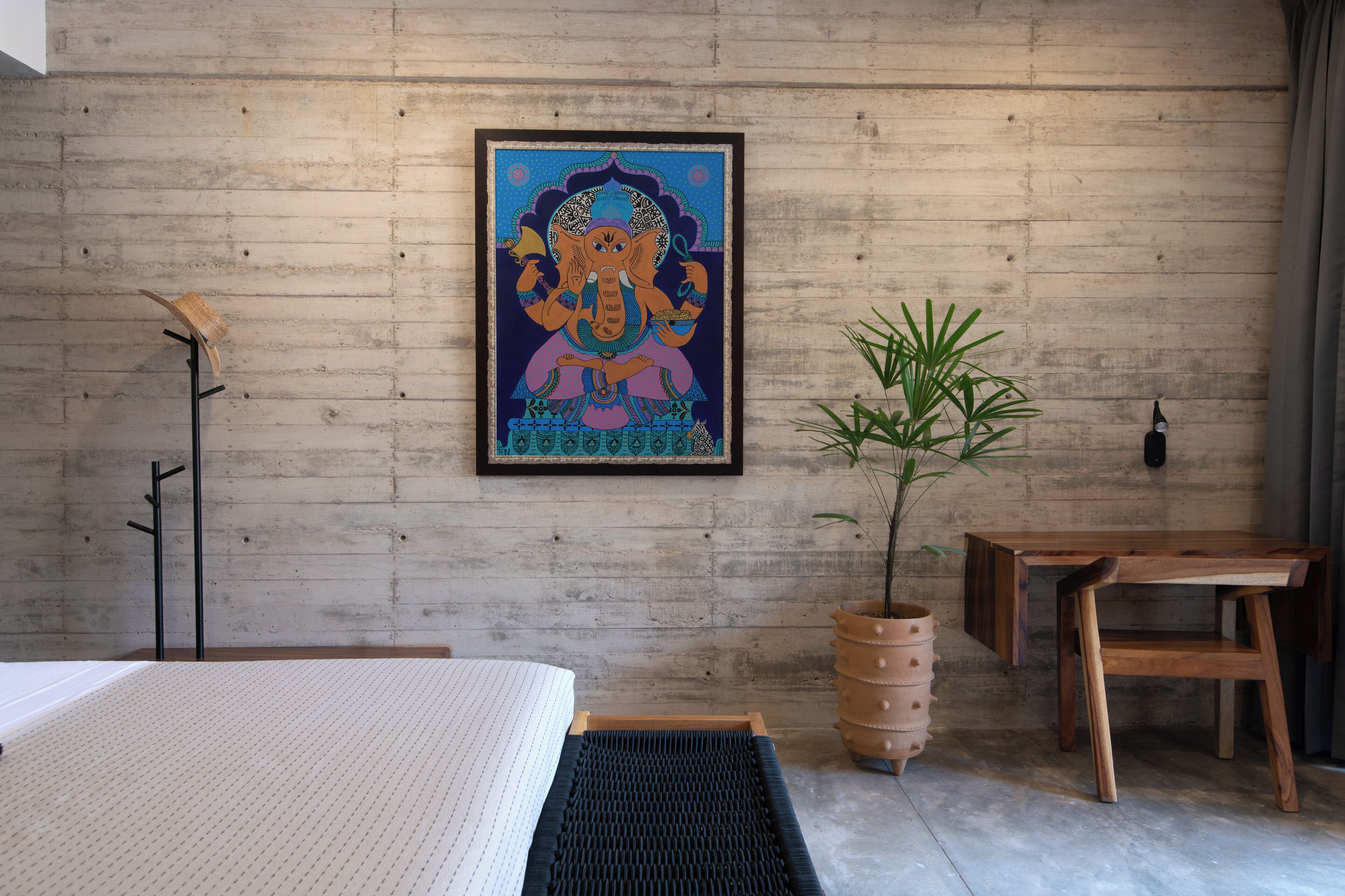 Ein Zimmer mit einem Bett, einem Holztisch, einer Topfpflanze und einem gerahmten Kunstwerk an der Wand.