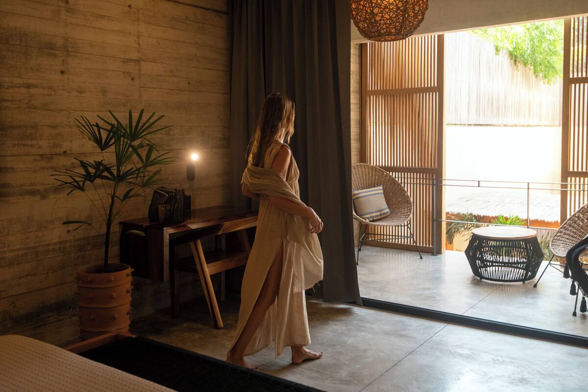 A woman in a white dress walking through a room with a wooden wall, a potted plant, and a view of an outdoor area with wicker furniture.