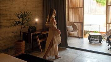A woman in a white dress walking through a room with a wooden wall, a potted plant, and a view of an outdoor area with wicker furniture.