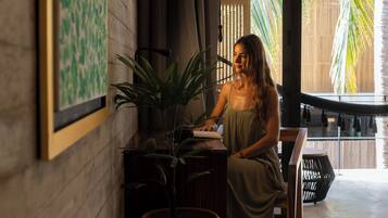 Una mujer sentada a una mesa de madera con un libro, rodeada de plantas en macetas y un ventanal con vistas a palmeras.