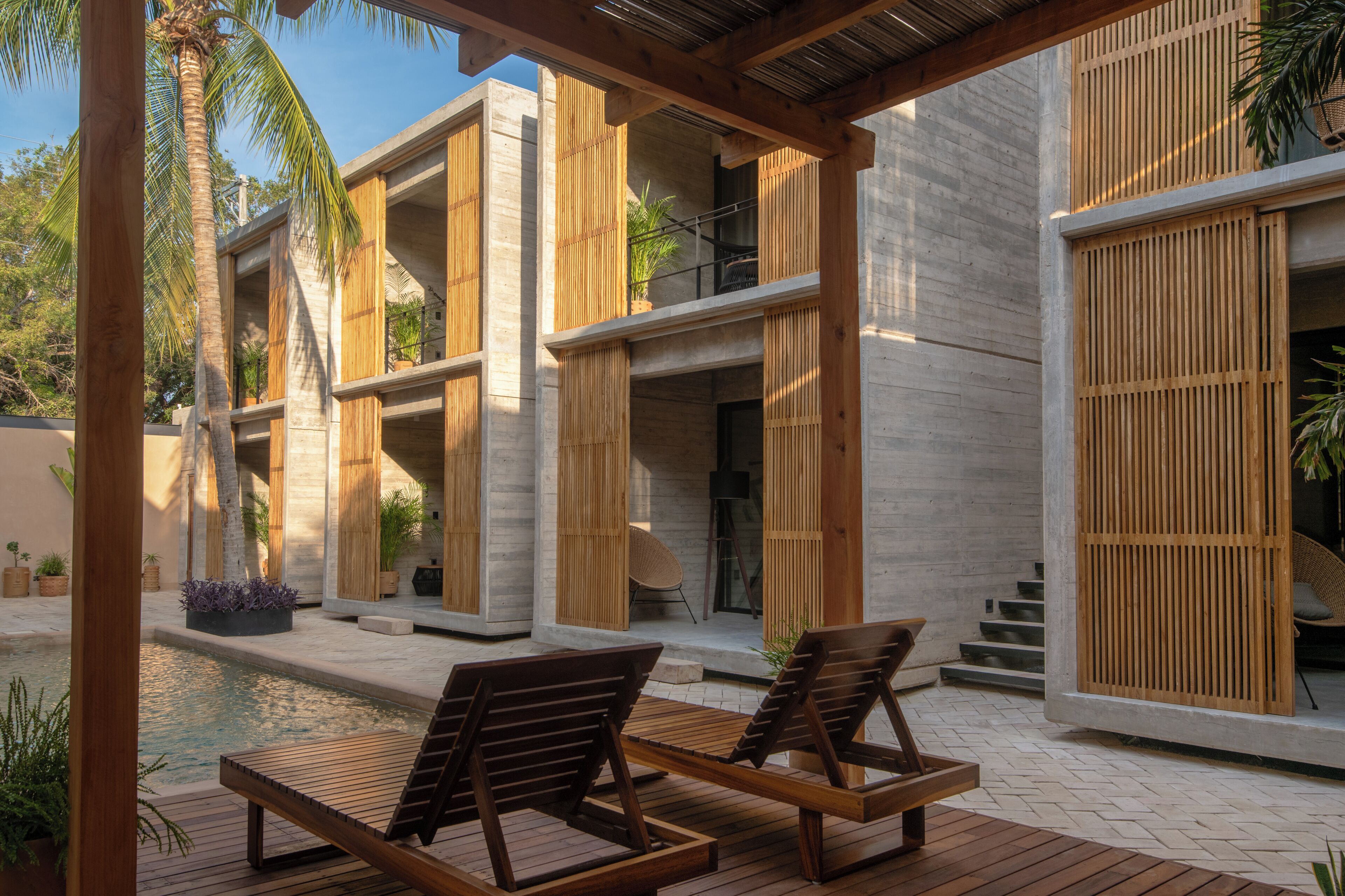 A modern outdoor pool area with wooden loungers, a concrete building with wooden slats, and a palm tree.