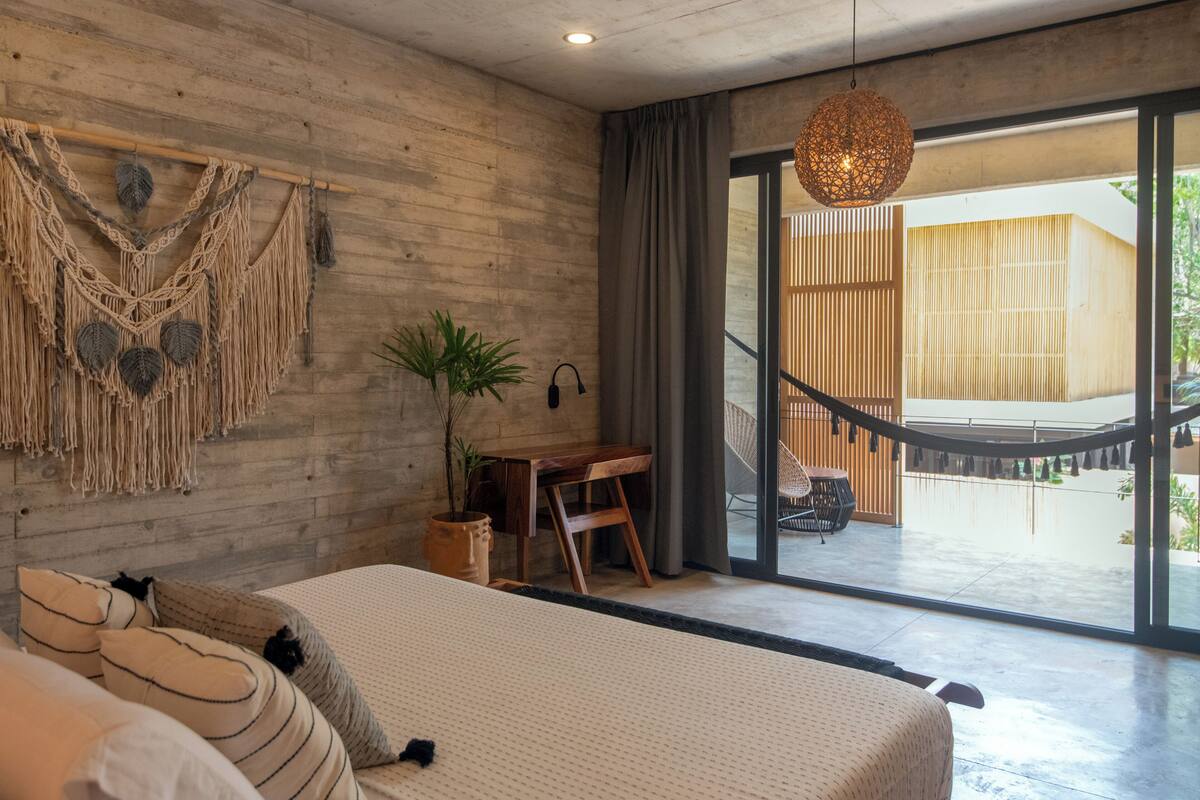 A modern bedroom with a large bed, a wooden desk, and a view of an outdoor area with a hammock and wooden structure.