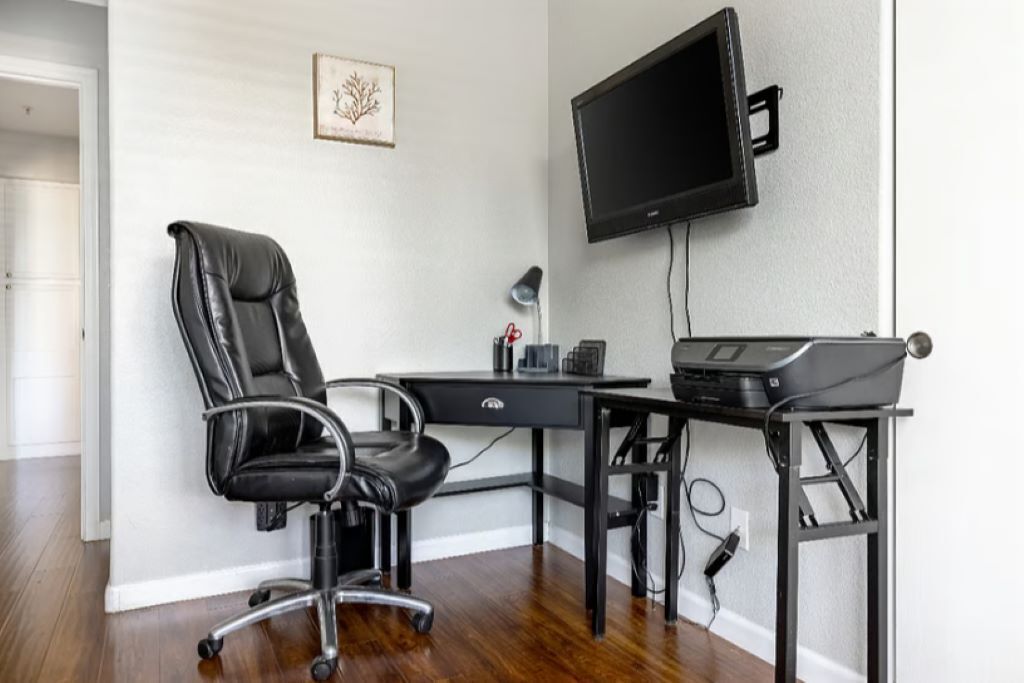 Oficina en esquina con sillón de cuero negro, escritorio con computadora e impresora, televisor de pantalla plana montado en la pared y un cuadro enmarcado.