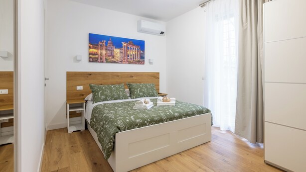 A bedroom with a bed, a nightstand, a wall-mounted air conditioner, a painting, and a wardrobe.