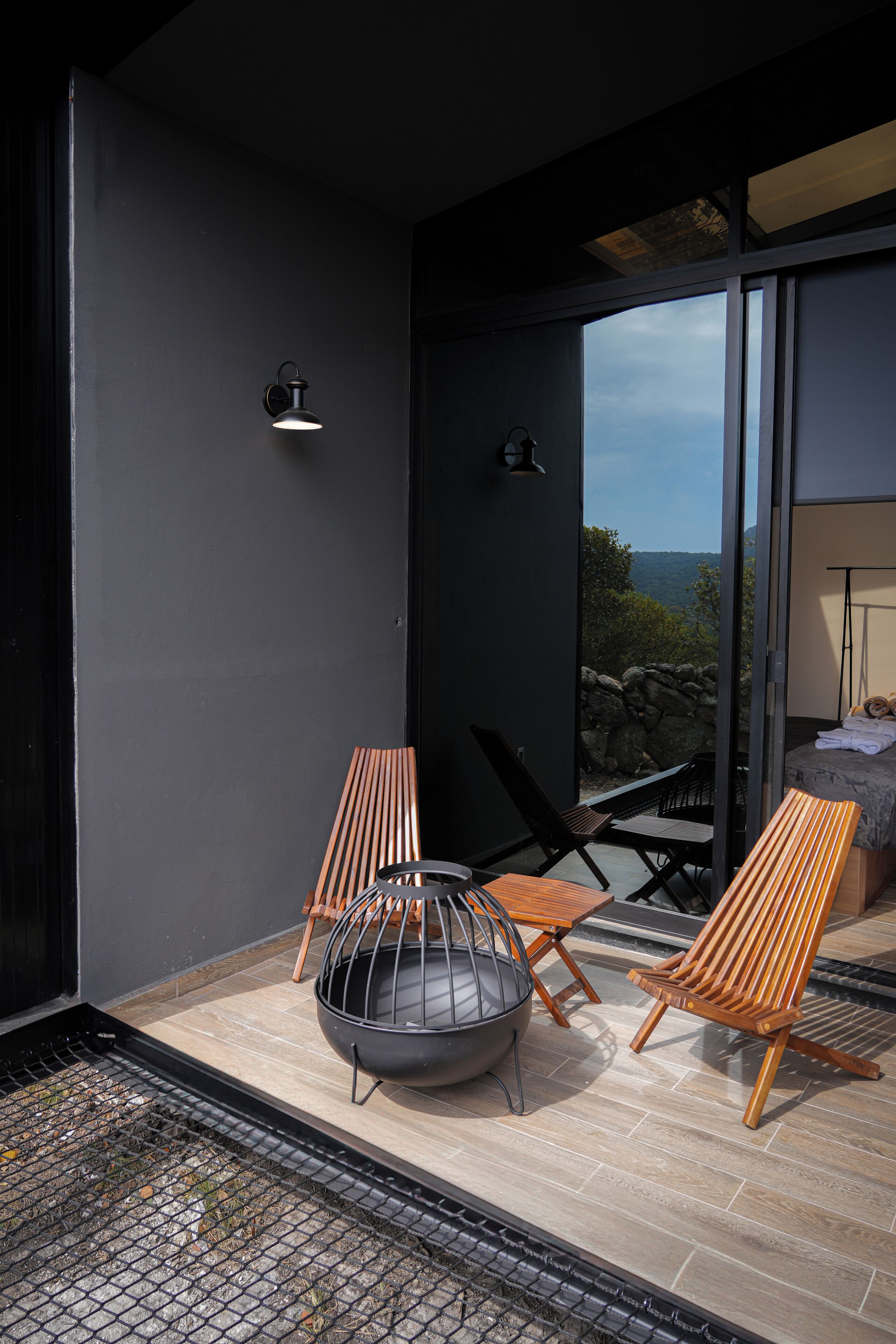 A modern outdoor seating area with two wooden chairs, a metal fire bowl, and a glass door leading to a bedroom.