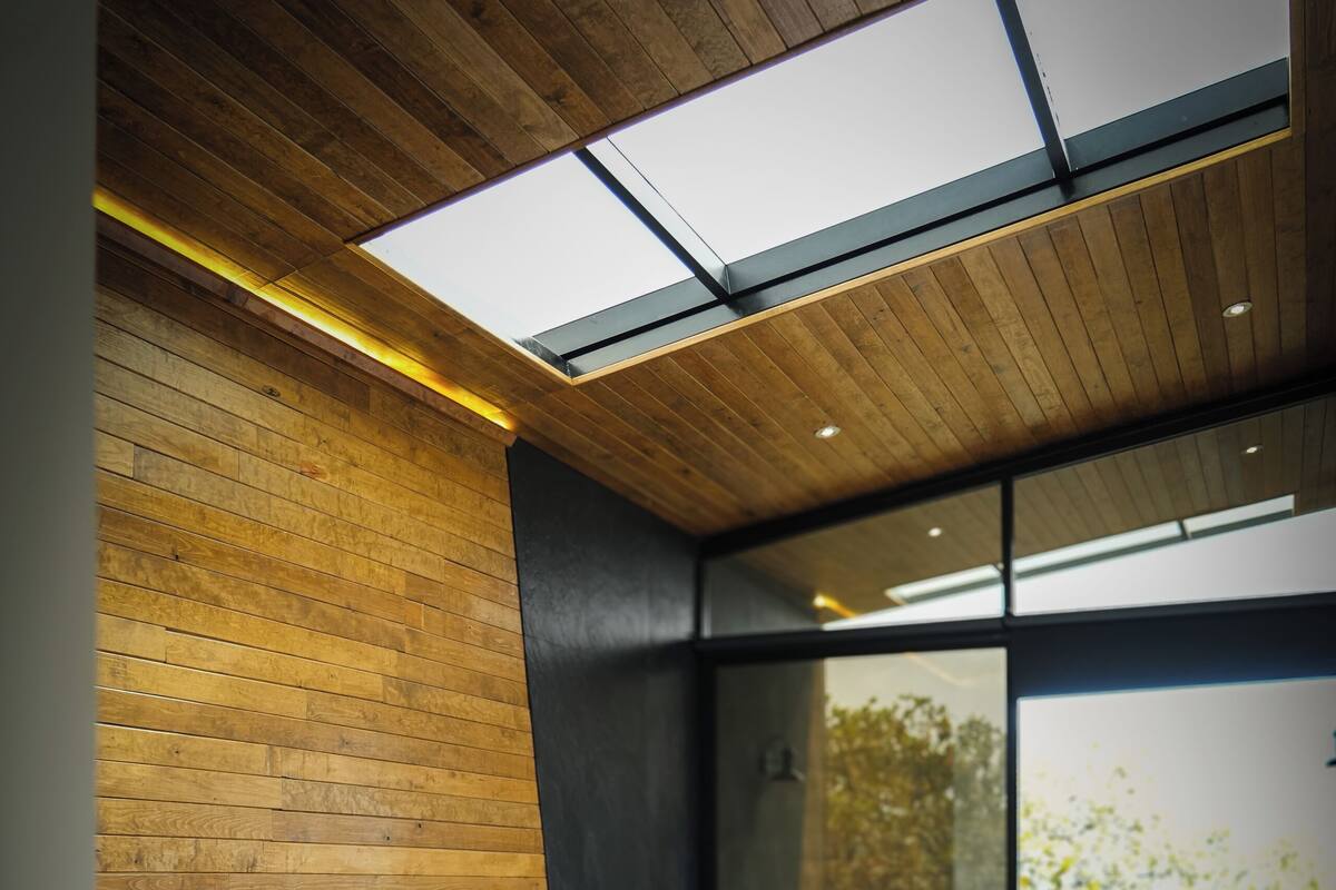 A modern bedroom with a wooden ceiling, a large skylight, a bathtub, and a view of trees.