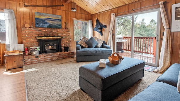 A cozy living room with a brick fireplace, a blue sofa, and a large window overlooking a deck. - Forest Edge Retreat (Big Bear City)