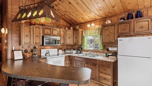 A wooden kitchen with a large island, bar stools, wooden cabinets, a refrigerator, and a window with curtains. - Forest Edge Retreat (Big Bear City)