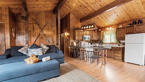 A cozy cabin interior with a blue sofa, wooden walls, a kitchen area, and a dining space. - Forest Edge Retreat (Big Bear City)
