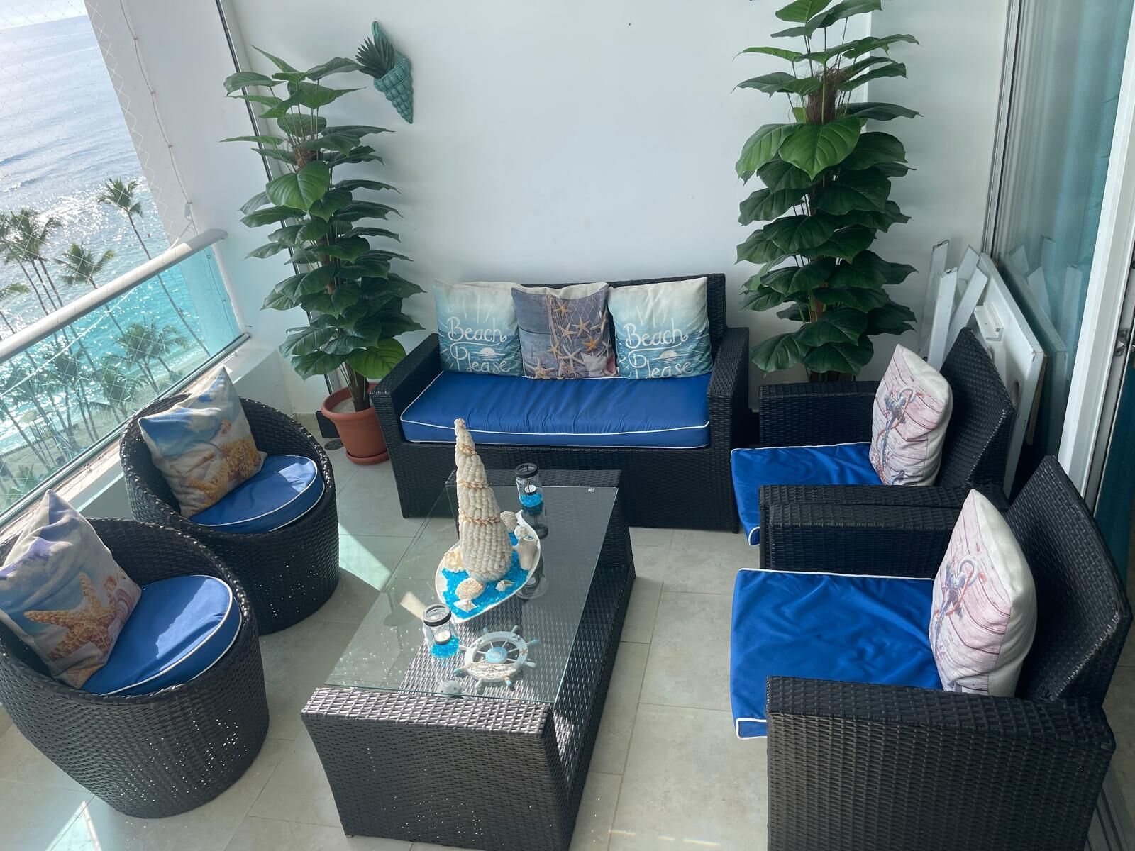 Un balcón con mobiliario de mimbre, un sofá con cojines azules y plantas en macetas.