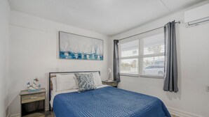 Une chambre à coucher avec un lit, une table de chevet, une fenêtre avec des rideaux, un climatiseur fixé au mur et un tableau au-dessus du lit.