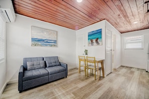 A room with a grey sofa, wooden dining table with chairs, a white door, and a ceiling with wooden beams.