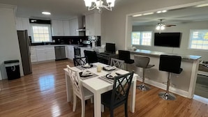 Une cuisine spacieuse avec des meubles blancs, un coin repas comprenant une table blanche et des chaises noires, et un téléviseur fixé au mur.