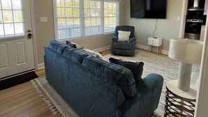 Un salon avec un canapé bleu, un ventilateur de plafond, un téléviseur à écran plat et un coin cuisine visible à travers une porte ouverte.