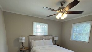 Une chambre à coucher avec un lit en bois, deux lampes de chevet, un ventilateur de plafond et deux fenêtres équipées de stores.