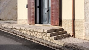 Ein Gebäude mit einer beigen Fassade, einem gepflasterten Eingangsbereich und einer Tür mit Glasscheibe und Vorhängen.