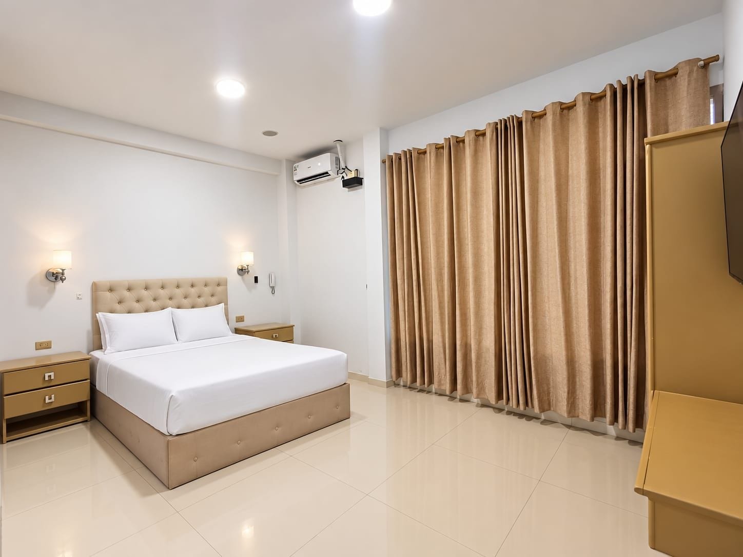 A hotel room with a bed, bedside tables, an air conditioning unit, a large window with curtains, and a wardrobe.