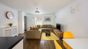 Ein modernes Wohnzimmer mit einer Couch, einem Couchtisch und einem Fernseher. Es gibt ein Fenster mit Jalousien, eine Pflanze und eine Uhr an der Wand.