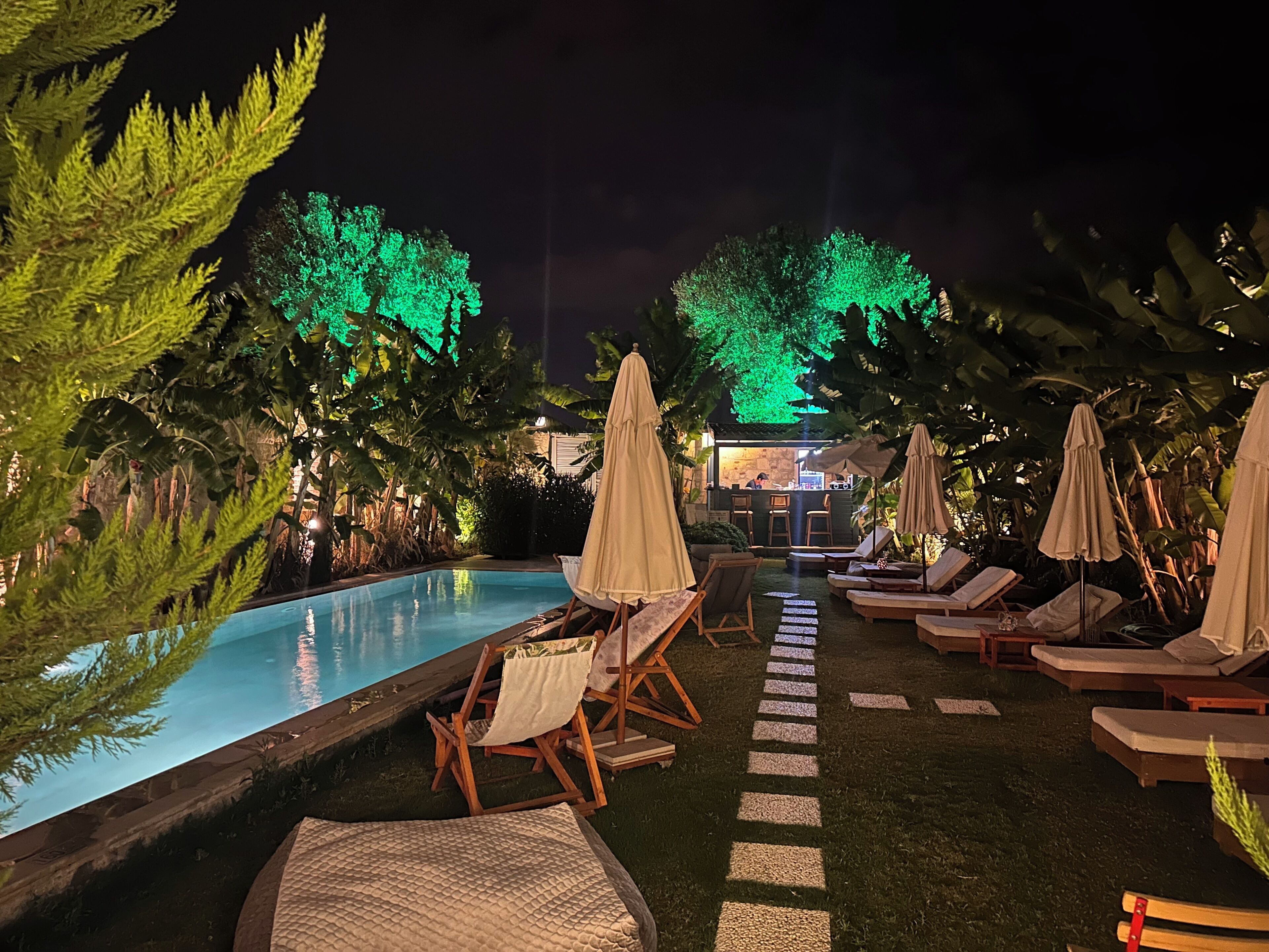 A nighttime pool area with lounge chairs and umbrellas, surrounded by trees.