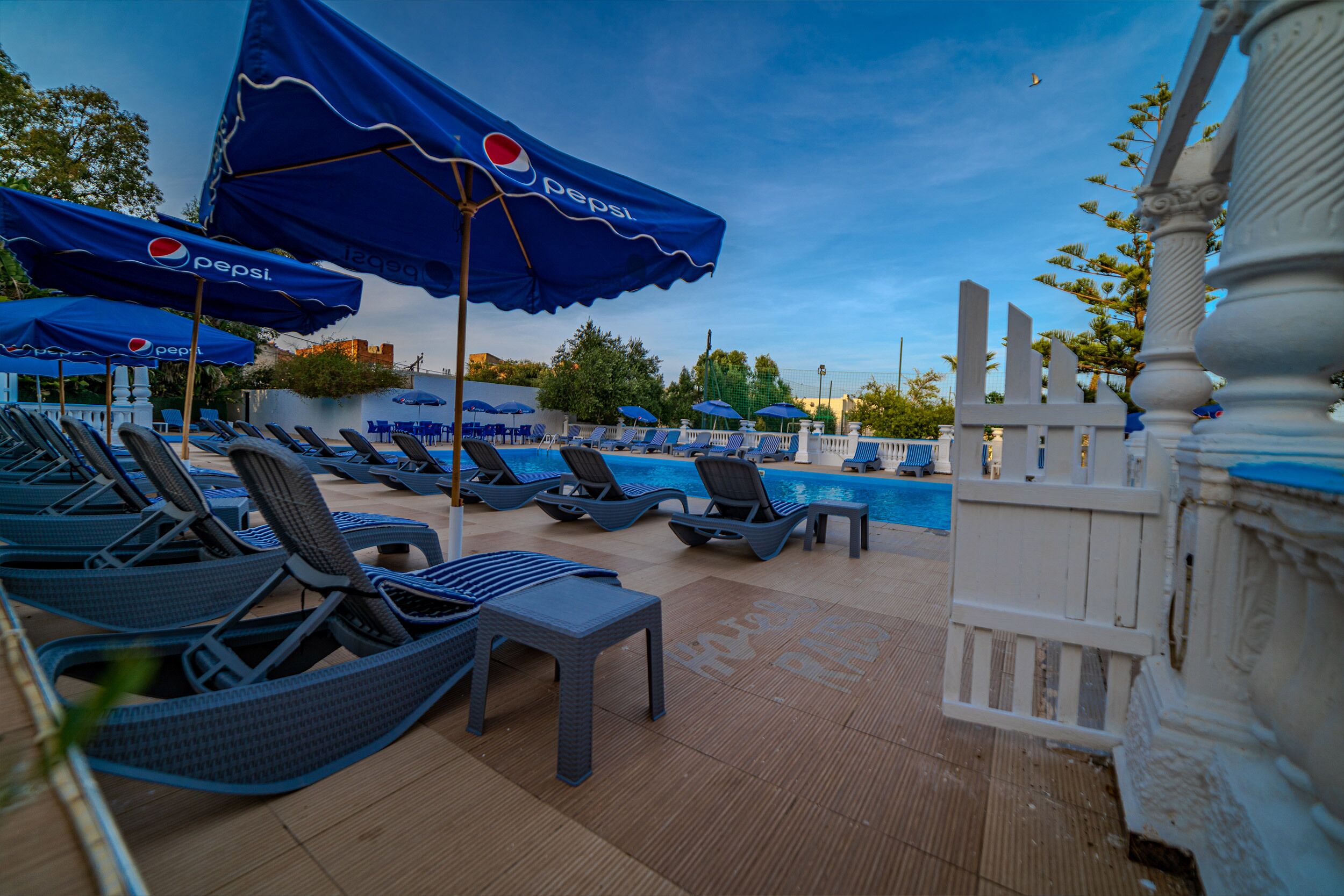 Zona de piscina con sillas de descanso y sombrillas, una piscina y una valla blanca.
