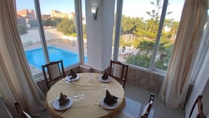 A dining area with a round table set for four, large windows with a view of a swimming pool and greenery, and curtains on the sides. - Rais El Marsa (Algiers)