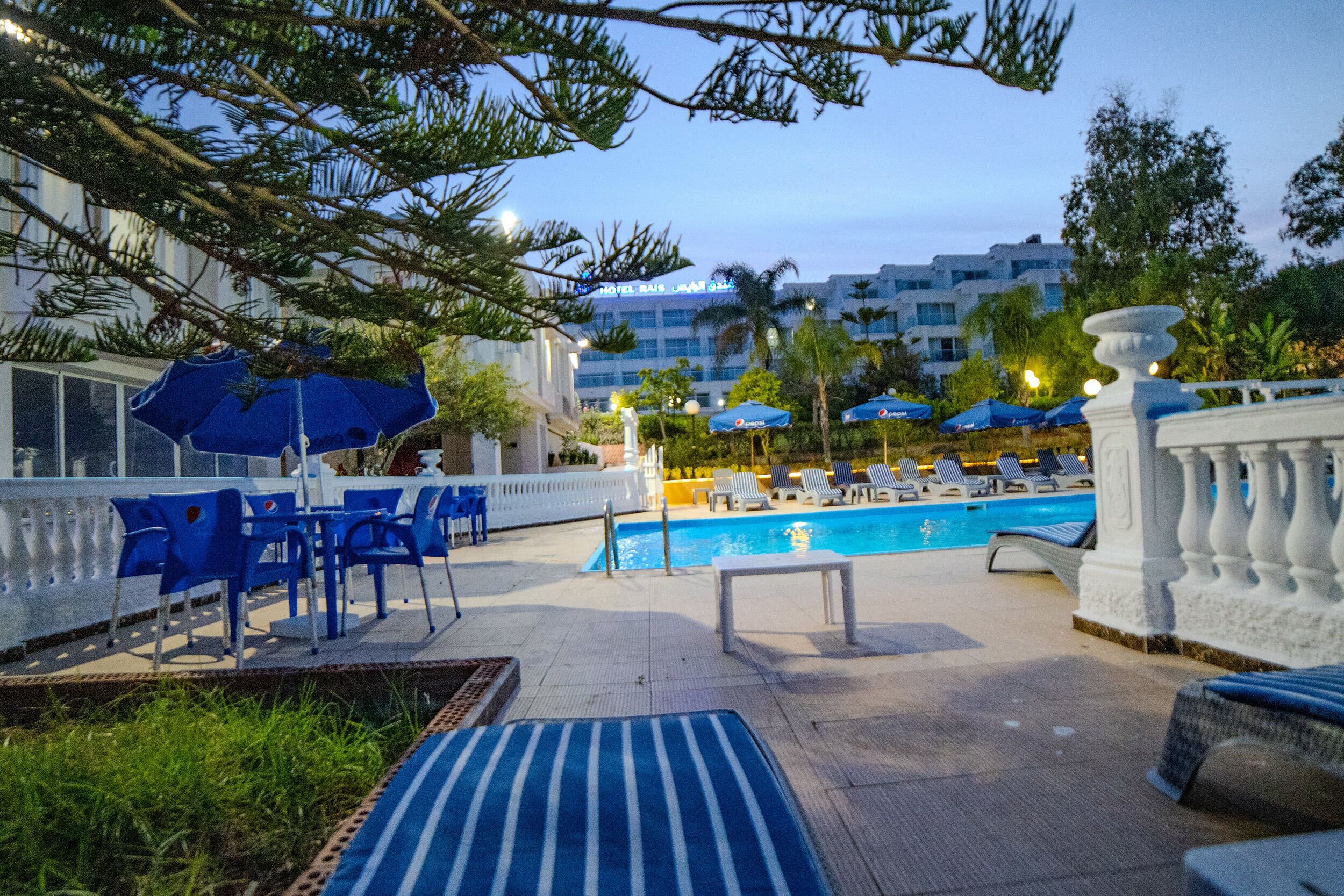 A poolside area with lounge chairs and umbrellas, a pool, and a hotel building in the background.