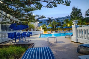 Ett område vid poolen med solstolar och parasoller, en pool och en hotellbyggnad i bakgrunden.