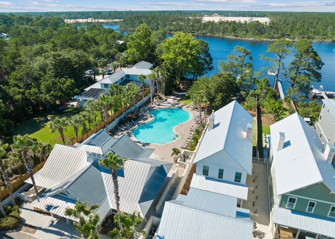 An aerial view of a resort with a central swimming pool, surrounded by multiple buildings with sloped roofs, likely guest rooms or villas, set against a backdrop of a lake and dense forest.