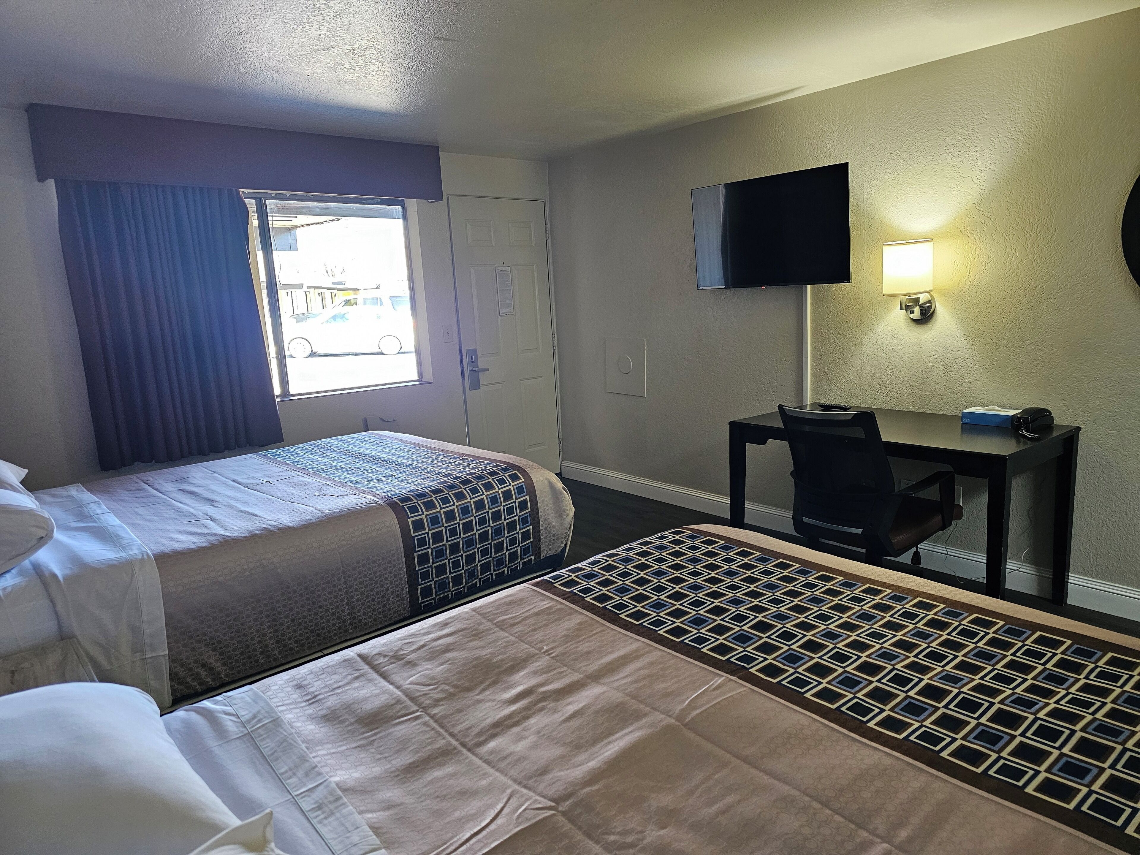 A hotel room with two beds, a desk, a TV, and a window with curtains.