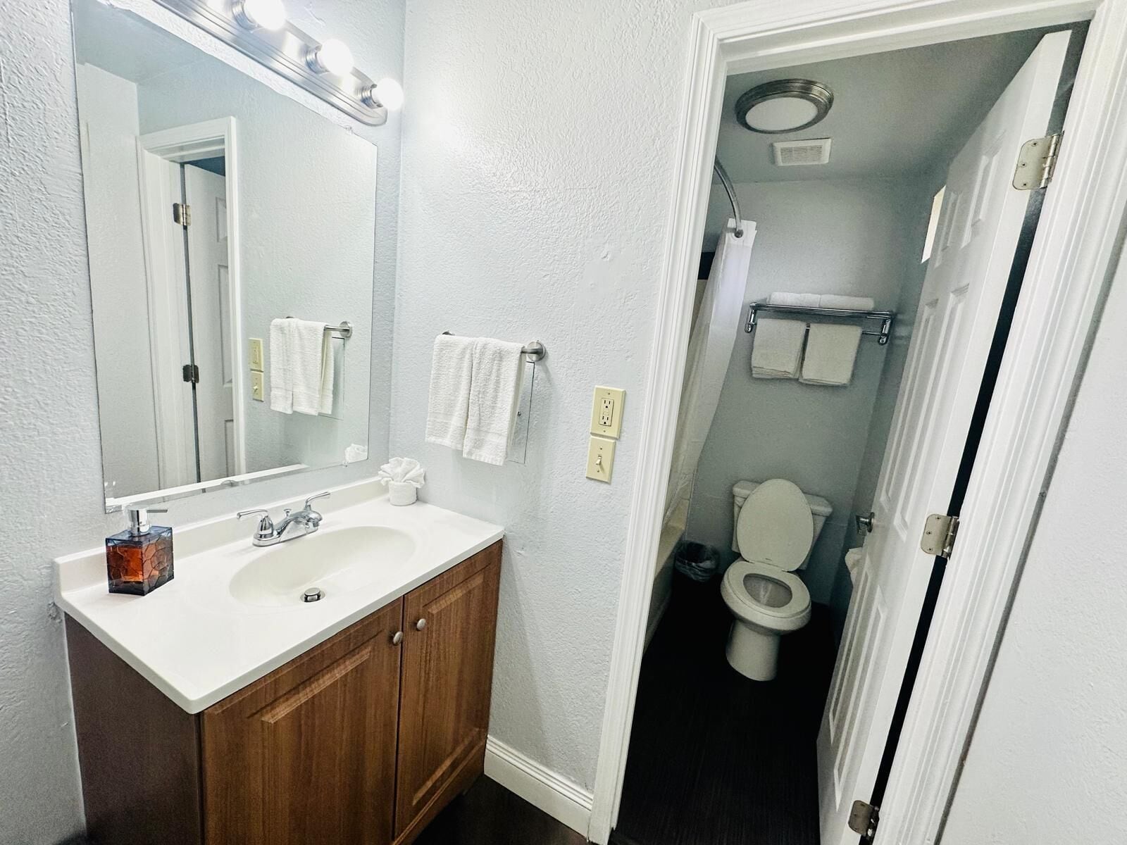 A bathroom with a white sink, wooden vanity, and a mirror with lights. There is a toilet visible through an open door.