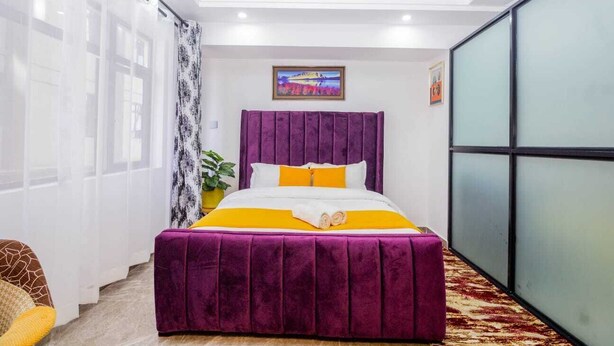 A bedroom with a purple bedspread, white and yellow pillows, and a painting on the wall. - Welcome to a spacious and cozy two bedroom @kilimani (Nairobi)