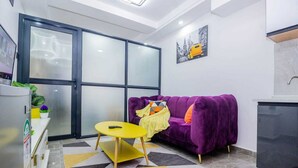A compact living space with a purple sofa, a small yellow table, a large window, and a kitchen area. - Welcome to a spacious and cozy two bedroom @kilimani (Nairobi)