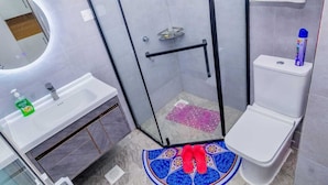 A bathroom with a glass-enclosed shower, a white toilet, a sink with a soap dispenser, and a patterned mat on the floor. - Welcome to a spacious and cozy two bedroom @kilimani (Nairobi)