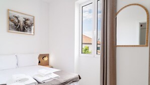 A bedroom with a bed, a bedside table, a wall-mounted lamp, a framed artwork, a window with a view, and a mirror.