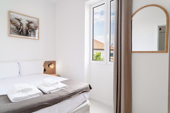 Une chambre à coucher comprenant un lit, une table de chevet, une lampe fixée au mur, un tableau encadré, une fenêtre donnant sur un paysage et un miroir.