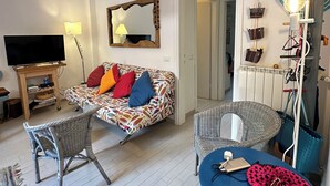 A living room with a sofa, a chair, a small table, a TV, a lamp, a mirror, and a wall-mounted shelf with bags.