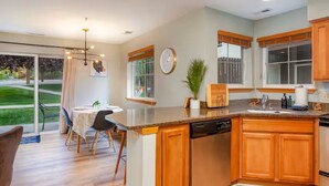 A modern kitchen with wooden cabinets, a dishwasher, and a dining area with a table and chairs. - GroundFloor+Pool+King Bed+HotTub+Skiing (Leavenworth)