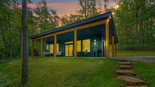 Eine moderne Hütte mit überdachter Terrasse, großen Fenstern und einem Steinweg, der zum Eingang führt.