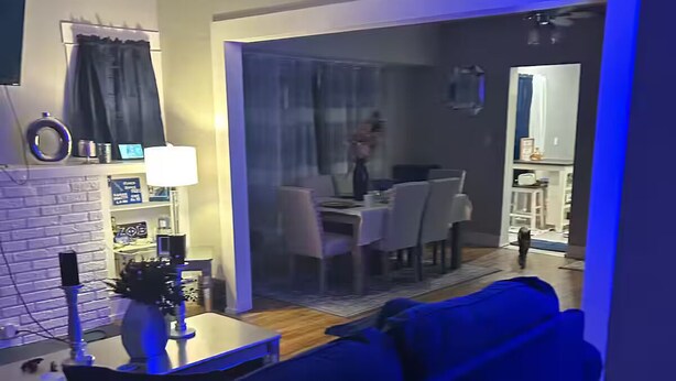 Una sala de estar con un sofá azul, un comedor con mesa y sillas, y una cocina visible a través de una puerta abierta.